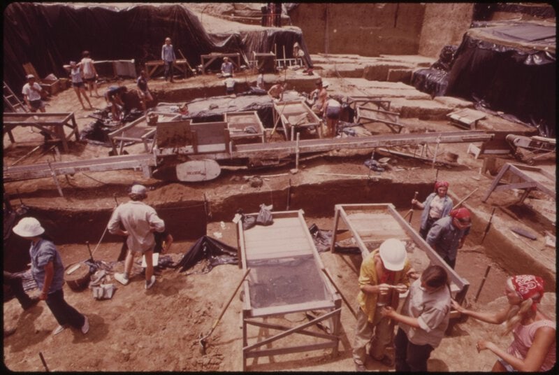 Shows a large active archaeological excavation with workers sieving and sorting soil, closely matching the section