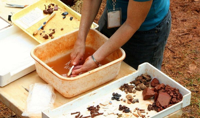 Shows hands-on archaeological fieldwork with sorted artifacts and cleaning in water, closely matching the section