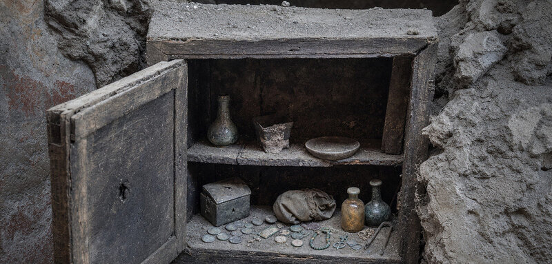 A figure opens a Pompeii cupboard sealed since AD 79, revealing coins, amulets, and perfume bottles preserved beneath…